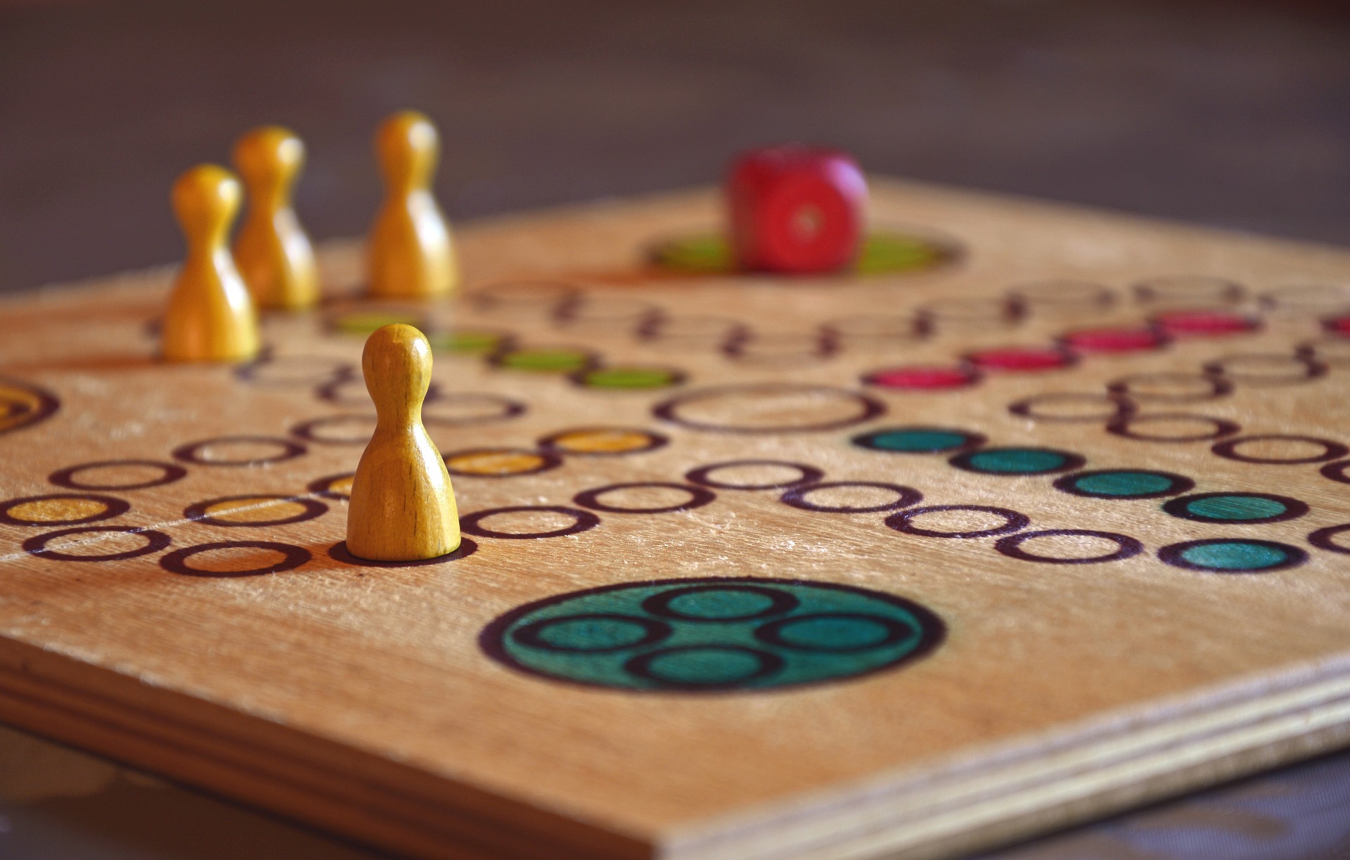 Gelbe Holzfigürchen und ein roter Würfel auf einem farbigen Spielbrett symbolisieren das Spiel „Mensch ärgere dich nicht“.