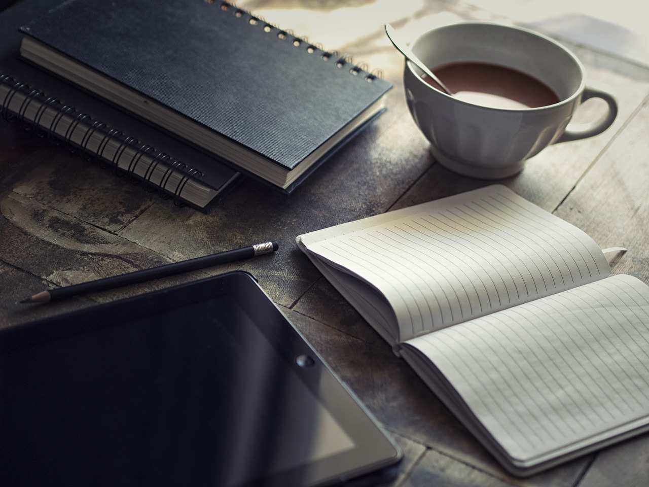 Kalender, Ipad, Notizbuch und Bleistift liegen bereit für unterschiedliche Notizen. Eine Tasse Kaffee macht die Schreibsituation gemütlich